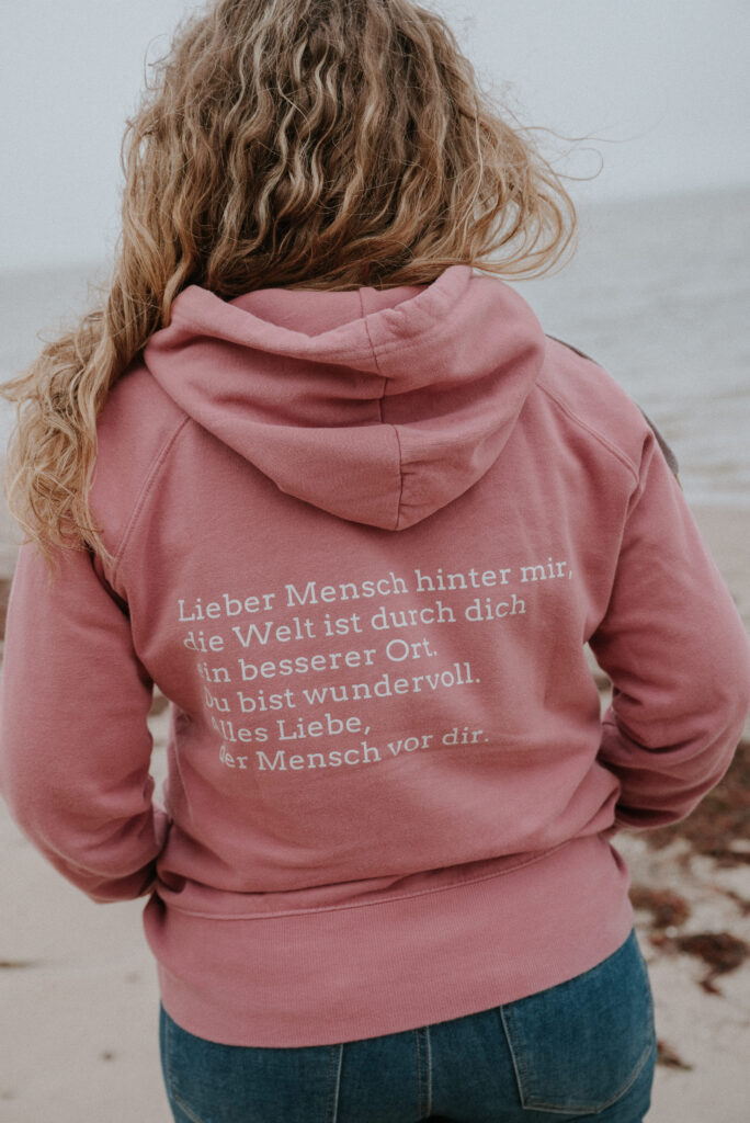 Claudia Hannemann am Strand mit Hoodie-Botschaft: Lieber Mensch hinter mir, die Welt ist durch dich ein besserer Ort – Wertschätzung für Fachkräfte in der Reflexintegration